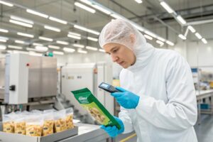 A quality control technician in protective gear inspecting a green packaged food pouch with a scanner in a clean production facility.