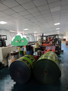 Factory interior with large rolls of printed laminated packaging film and production equipment.
