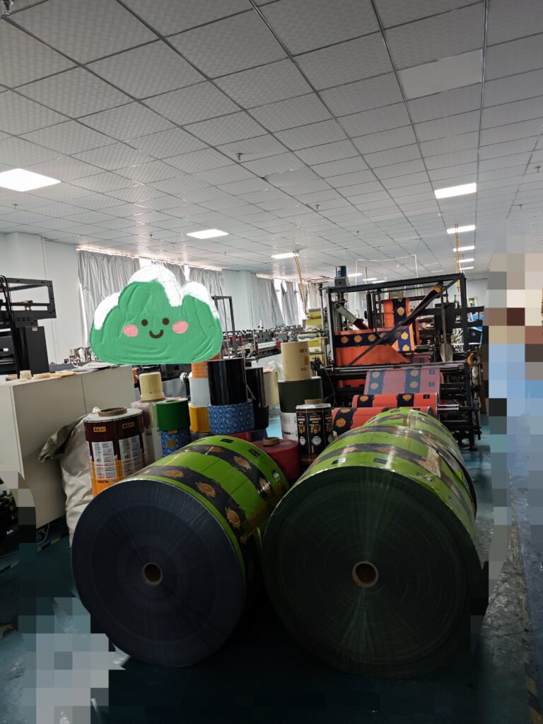 Factory interior with large rolls of printed laminated packaging film and production equipment.